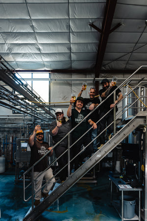 Paradox Brewery Brewers standing on the stairs in the brewhouse holding up a beer to cheers.