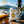 A can of Pilsner Light beer by Paradox Brewing stands on a wooden dock in a lakeside setting, with a cyclist blurred in the background, sunglasses, a helmet, and a paddleboard visible in the foreground. A yellow circle badge in the top left reads '100 CALORIES'