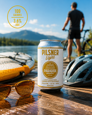 A can of Pilsner Light beer by Paradox Brewing stands on a wooden dock in a lakeside setting, with a cyclist blurred in the background, sunglasses, a helmet, and a paddleboard visible in the foreground. A yellow circle badge in the top left reads '100 CALORIES'