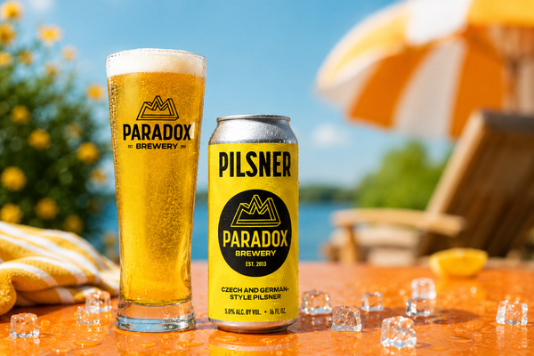 A glass and can of Paradox Brewery Pilsner beer on an outdoor table with ice cubes, a striped towel, and a beach umbrella in the background.